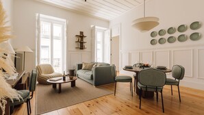 Ein geräumiges Wohnzimmer mit einer Couch, Sesseln, einem Couchtisch und einem Essbereich mit Tisch und Stühlen. Große Fenster sorgen für viel Tageslicht, und eine Deckenleuchte erhellt den Raum.
