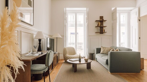 Ein modernes Wohnzimmer mit Sofa, Sessel und Couchtisch. Es gibt einen Schreibtisch mit Lampe und Stuhl. Der Raum verfügt über große Fenster und einen Balkon.