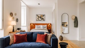 A modern bedroom with a bed, a blue sofa with red cushions, a bedside table with a lamp, a wall-mounted mirror, and a framed abstract painting. - Ando Living - Corpo Santo House - 202 (Lisboa)