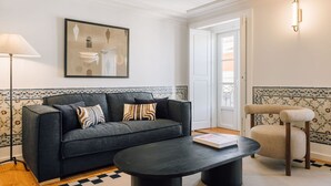 A living room with a dark grey sofa, patterned cushions, a black coffee table, and a beige armchair. A framed abstract artwork hangs on the wall, and a floor lamp is visible. - Ando Living - Madalena House - 501 (Lisboa)