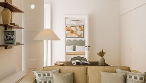 A modern living room with a sofa, a coffee table, and a lamp. There is a door leading to another room with a bed and a painting on the wall. - Ando Living - Flores 25 House - 302 (Encarnação)
