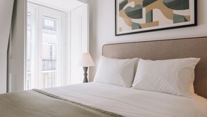 Habitación de hotel con cama, mesita de noche, lámpara y ventana con vistas a edificios.
