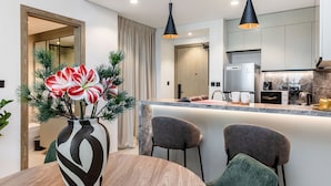 A modern kitchen with a built-in oven, a coffee machine, and a refrigerator. There is a dining area with a wooden table and chairs, and a vase with flowers on the table. - Stylish 1BR Apartment at Golf Gate (Dubai)
