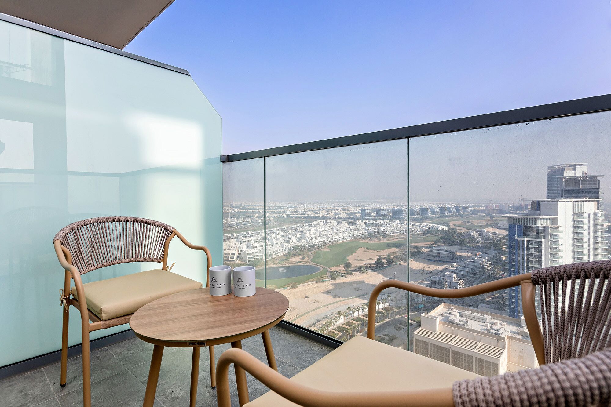 A balcony with two wicker chairs and a small round table, a glass partition, and a city view.