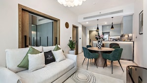 A modern living room with a sofa, a dining area with a round table and chairs, and a kitchen in the background. - Stylish 1BR Apartment at Golf Gate (Dubai)