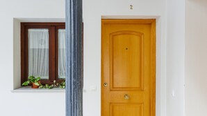 Una puerta de madera con el número '2' encima, una ventana con cortinas y una planta en maceta, y una pared blanca.