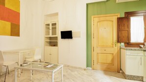 Una habitación con una mesa de vidrio, sillas blancas, una puerta de madera, un televisor montado en la pared y una zona de cocina con armarios.