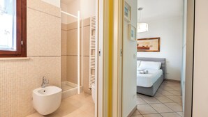 Un baño con lavamanos, ducha e inodoro. En la habitación contigua hay una cama.