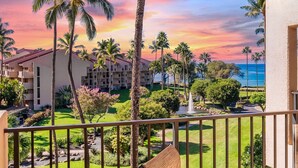 A hotel with a view of a landscaped garden, palm trees, and a building with multiple balconies. - NICELY UPDATED-OCEAN VIEWS-PRIME LOCATION-LARGE LANAI-AC-WELL EQUIPPED (Kihei)