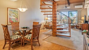 A spacious living area with a wooden spiral staircase, a glass dining table with floral chairs, and a large window offering a view of palm trees. - NICELY UPDATED-OCEAN VIEWS-PRIME LOCATION-LARGE LANAI-AC-WELL EQUIPPED (Kihei)