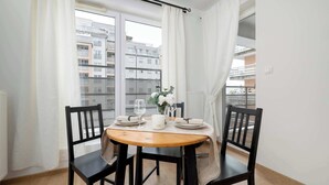 A dining area with a wooden table and black chairs, a vase with flowers, and a view of buildings through large windows. - Air-conditioned Apartment | Balcony, Parking and Workspace (Kraków)