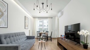 A modern living room with a grey sofa, a dining table with chairs, a TV on a wooden cabinet, and a chandelier. - Air-conditioned Apartment | Balcony, Parking and Workspace (Kraków)