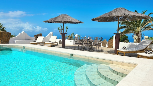 Ein klares, blaues Schwimmbecken mit Liegestühlen und Sonnenschirmen, ein Essbereich mit Tisch und Stühlen sowie Blick auf den Ozean.