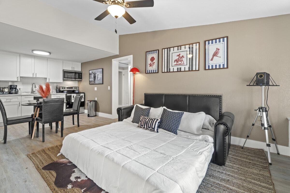 A bedroom with a bed, a desk, and a chair, a kitchenette, and a ceiling fan.