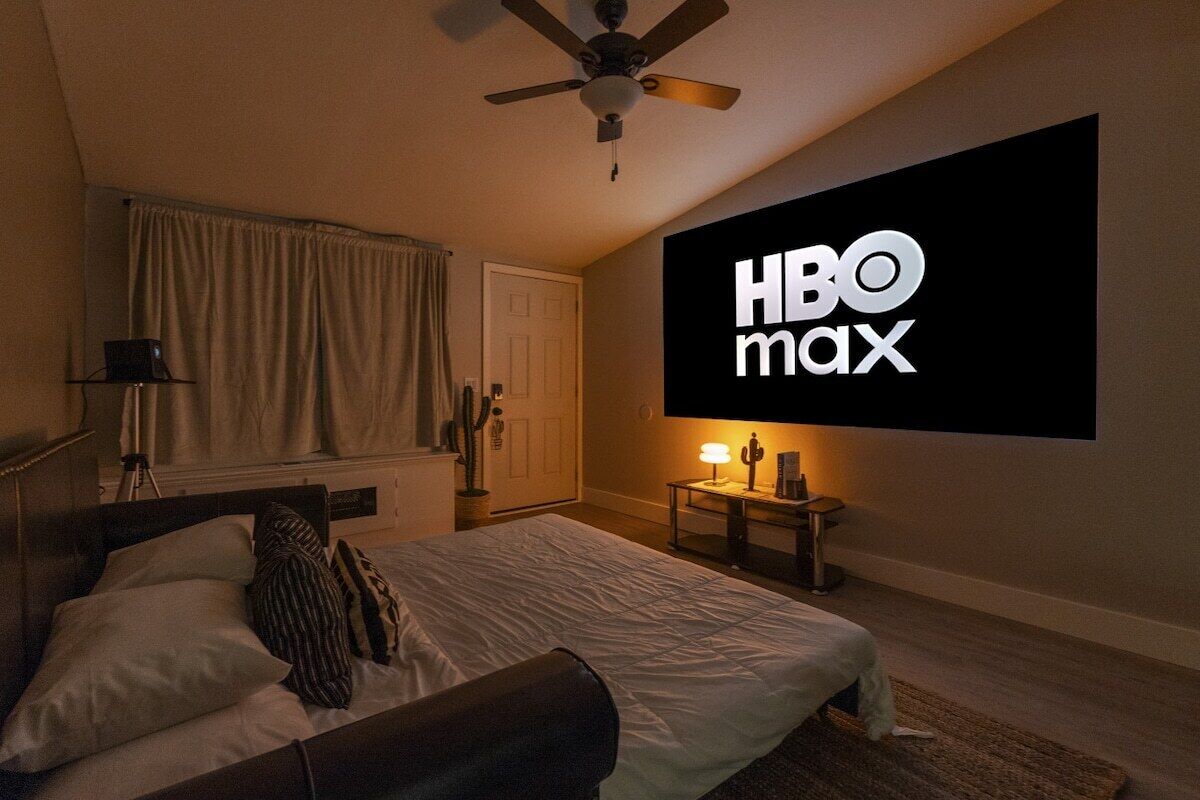 Une chambre avec un lit, un ventilateur de plafond, un téléviseur fixé au mur diffusant HBO Max, et une table de chevet avec une lampe.