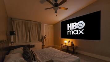 Une chambre avec un lit, un ventilateur de plafond, un téléviseur fixé au mur diffusant HBO Max, et une table de chevet avec une lampe.