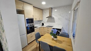 Et lille køkken med træskabe, indbygget ovn, mikrobølgeovn og komfur. Der er et spisebord med stole og en potteplante.