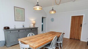 A dining area with a wooden table and white chairs, a grey cabinet, and two hanging lamps. - Braeburn (Bamburgh)