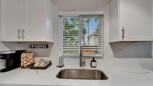 A modern kitchen with white cabinets, a coffee maker, a basket of coffee pods, and a window with blinds. - Private Hot Tub - Close to Beaches - Fun Game Room (Clearwater)