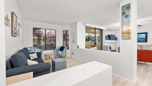 Una sala de estar moderna con un sofá gris, un sillón azul y una mesa de centro blanca. Hay un ventanal con persianas, un televisor montado en la pared y un cuadro sobre el sofá.