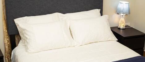 Una cama con ropa de cama blanca y un cabecero oscuro, una lámpara de noche y una mesita de noche oscura.