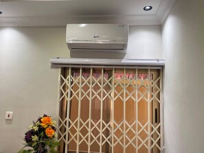 Une pièce dotée d’un climatiseur fixé au plafond, d’une porte coulissante ornée d’un motif en treillis et d’un arrangement floral posé sur le sol.