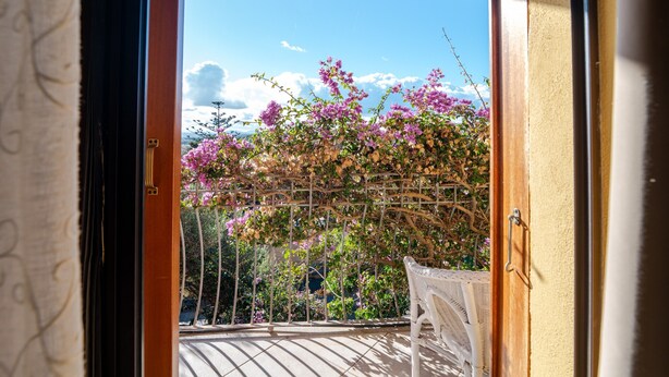 A view through a wooden-framed window with a metal gate, leading to a balcony with a white chair and a fan. - Room 'La Chicca Di Francesca- Room 3' with Private Terrace, Wi-Fi and Air Conditioning (Santa Teresa Gallura)