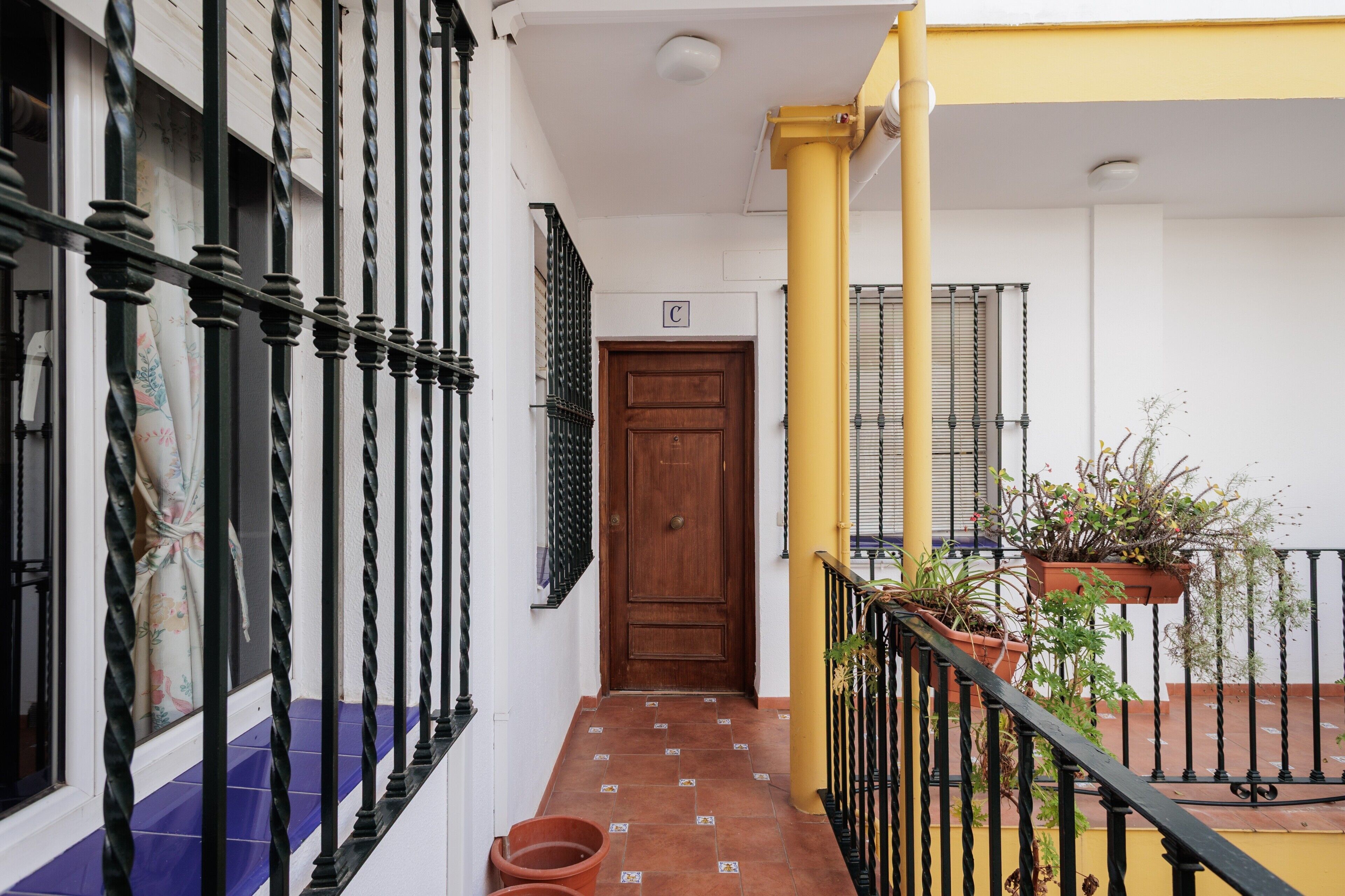 Un couloir avec une porte en bois, un mur jaune, une rambarde noire et des plantes en pot.