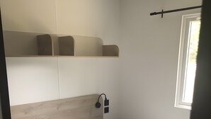 Una cama individual con ropa de cama blanca y gris, un cabecero de madera, una mesita de noche con lámpara y una ventana con luz natural.