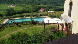 Una piscina con sillas de descanso, un edificio con un ventanal arqueado y vistas a colinas verdes.