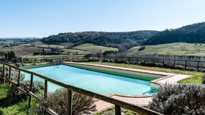 Una piscina rectangular con muelle de madera y valla de metal, rodeada de césped y colinas al fondo.
