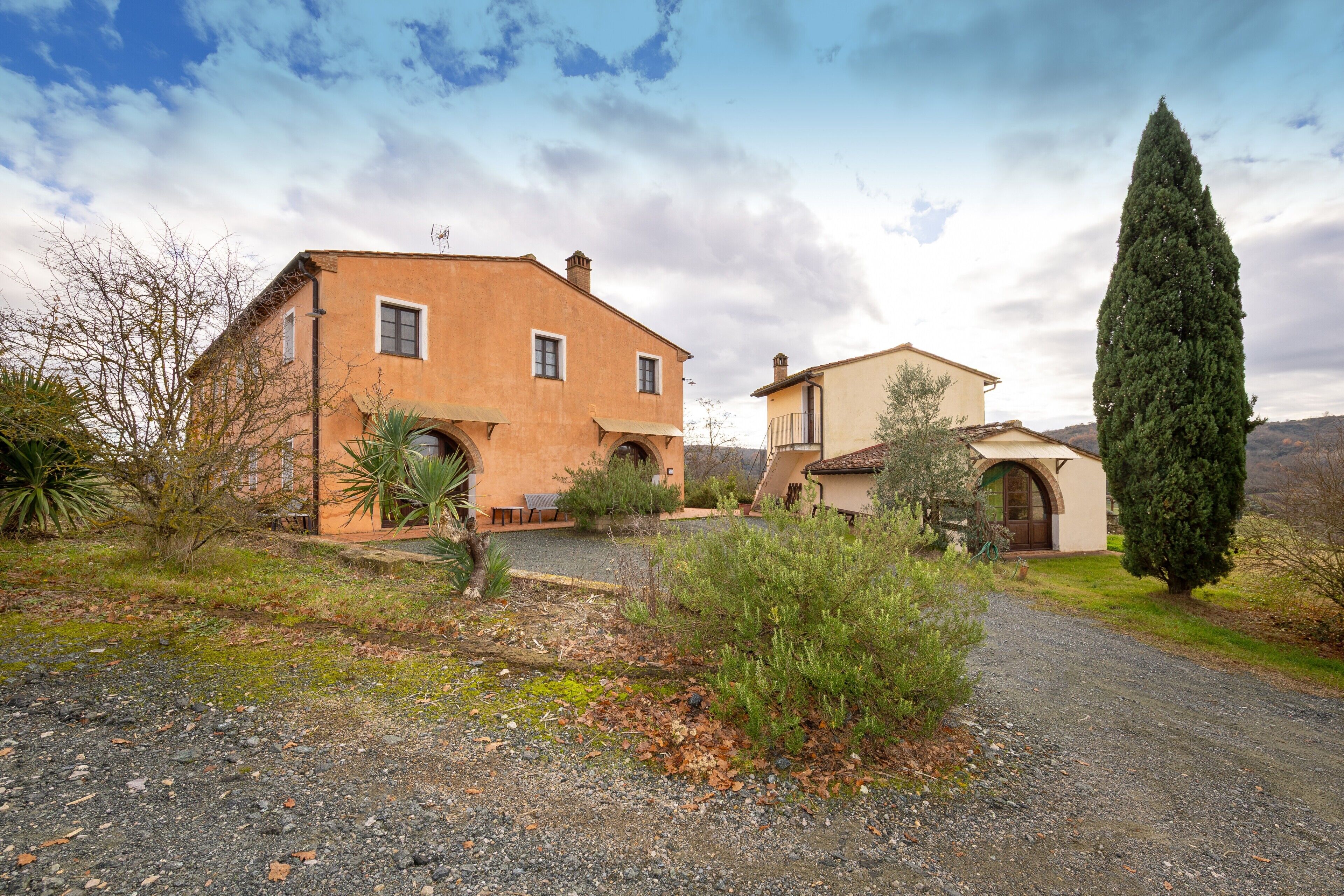 A two-story house with a gravel driveway, a tall cypress tree, and a clear sky.