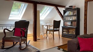 A loft-style living room with a high ceiling, wooden beams, a glass coffee table, a sofa, a rocking chair, a bookshelf, and a dining area with a table and chairs.
