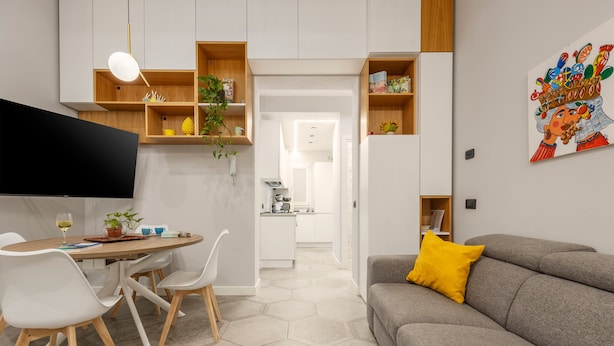 A modern living room with a grey sofa, a dining table with chairs, a flat-screen TV, and a kitchen in the background.