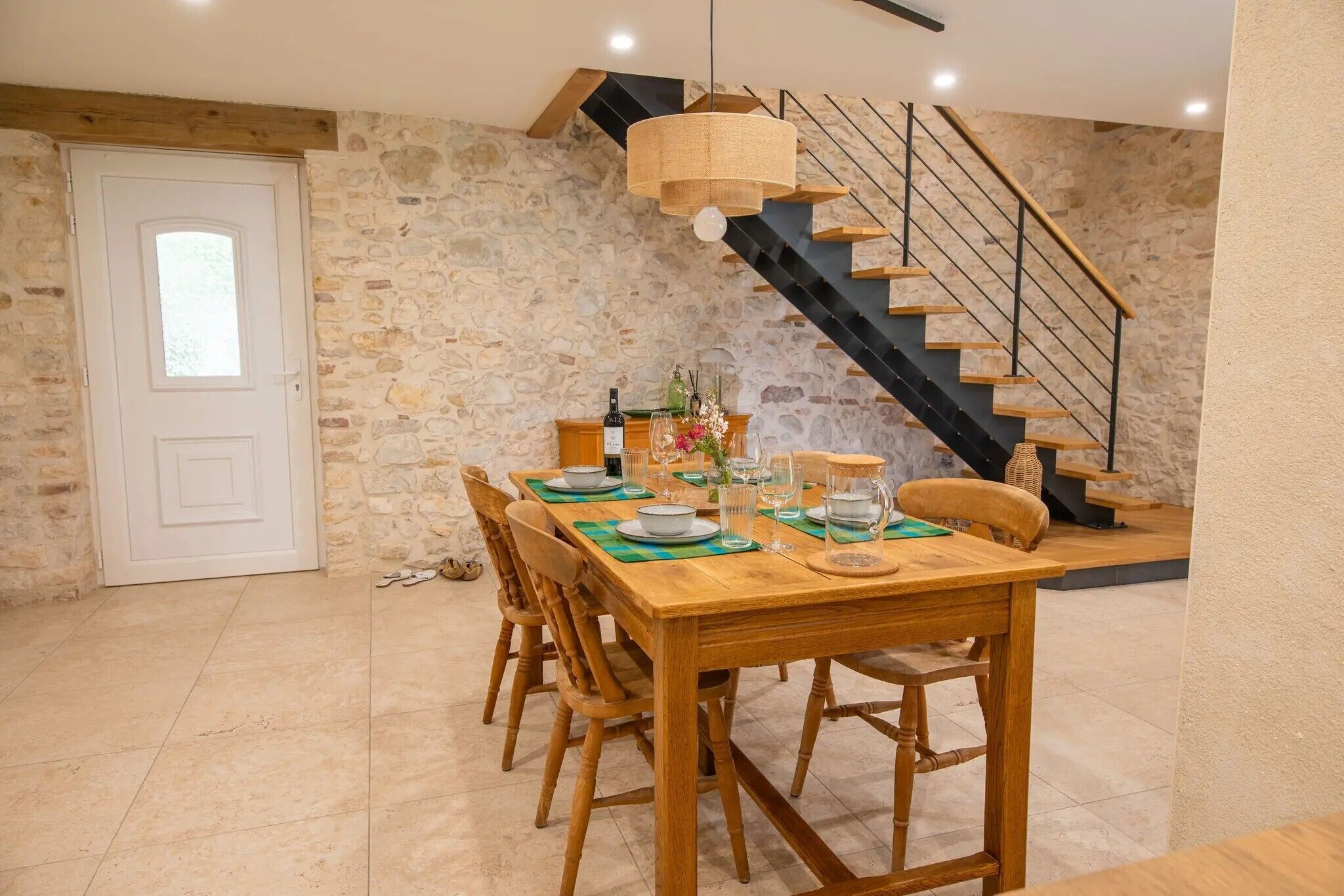 Un coin repas avec une table en bois dressée pour six personnes, un mur en pierre, un escalier et une porte blanche.