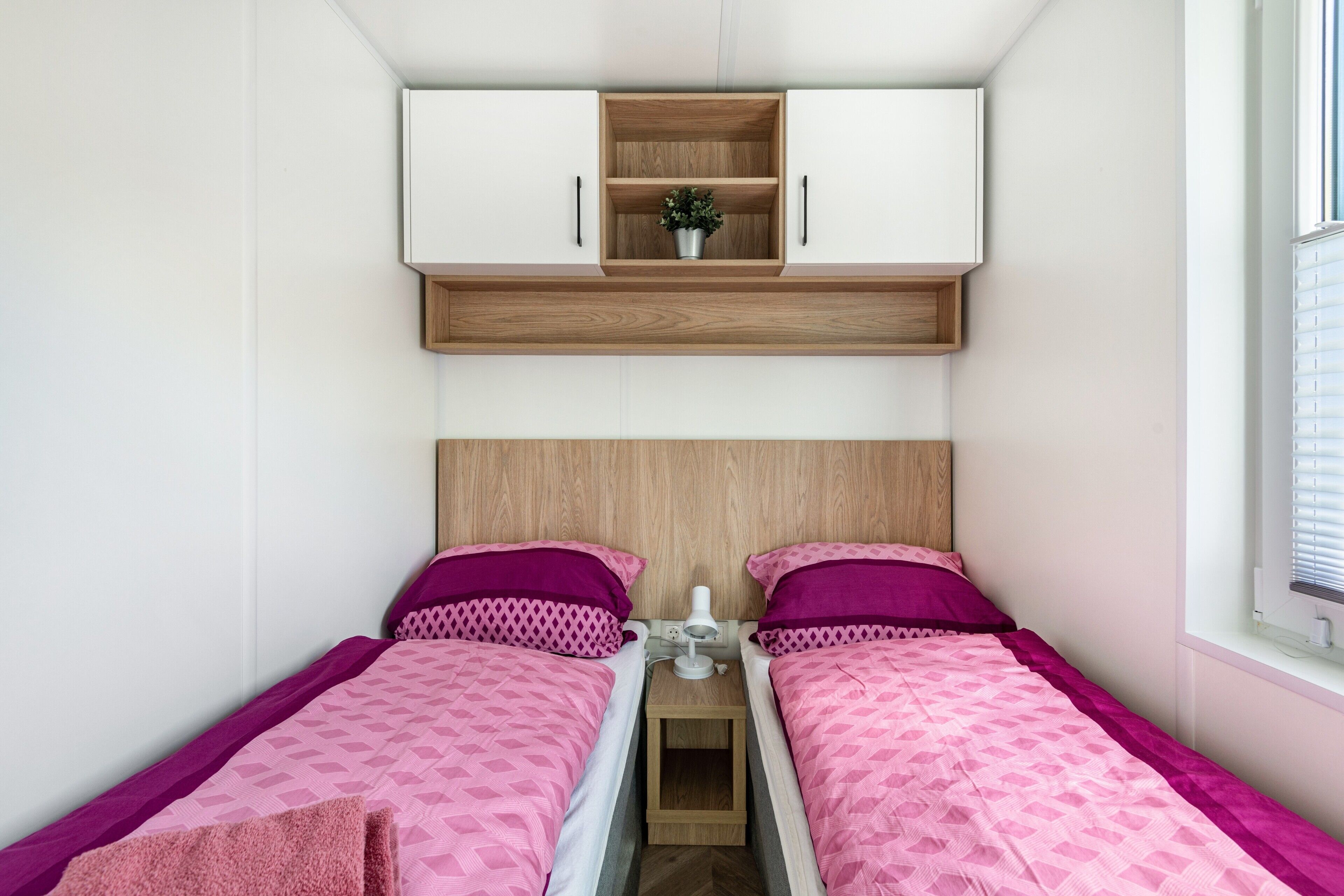Een kamer met twee eenpersoonsbedden, een klein nachtkastje, een witte kast boven de bedden en een raam aan de rechterkant.