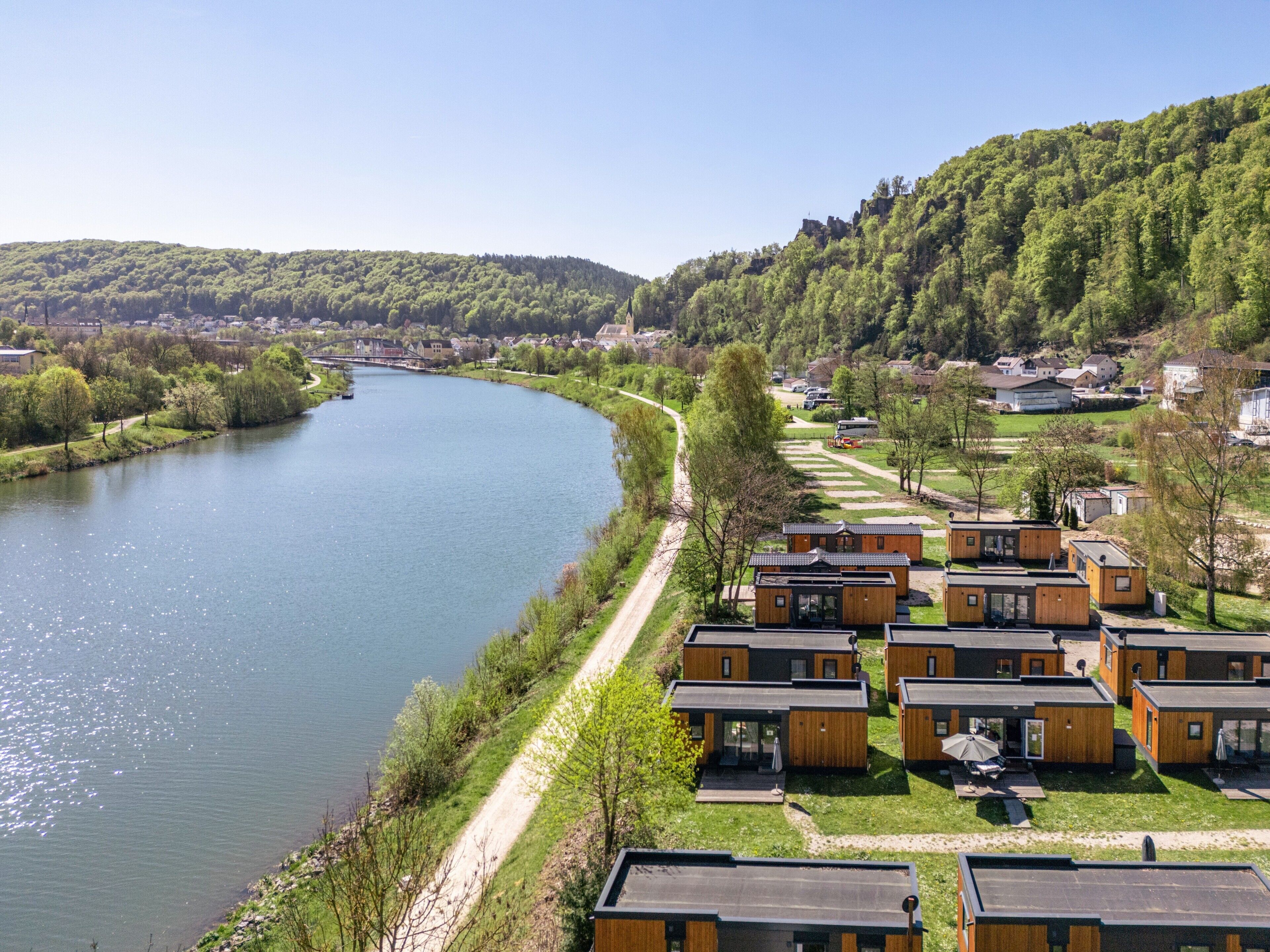 Blick auf einen Fluss mit einem Hotel und modernen Holzhäusern, wolkenlosem Himmel und umliegender Vegetation.