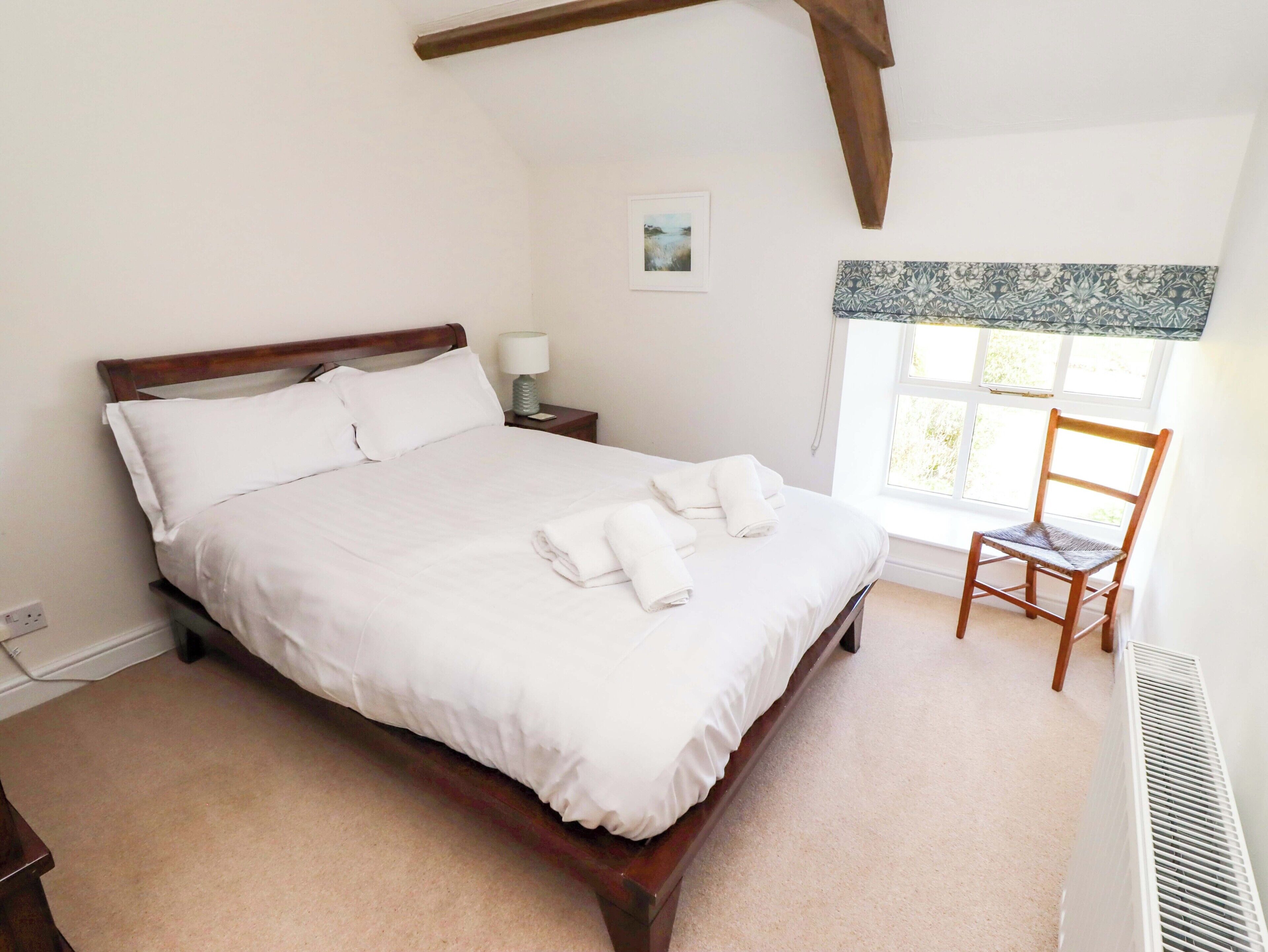 A neatly made bed with white linens, a wooden headboard, a bedside table with a lamp, a chair, and a window with a view of greenery.