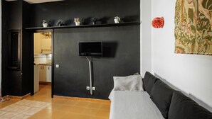 A room with a black wall, a flat-screen TV mounted on it, a sofa with a cushion, and a door leading to a kitchen.