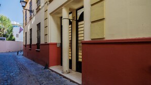 A narrow cobblestone street with a building featuring a red and beige facade, a black gate, and a flag.