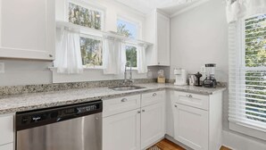 A modern kitchen with white cabinetry, a granite countertop, a dishwasher, and a window with curtains. - The Pearl, 2 Unit Stay for 10 (St. Augustine)