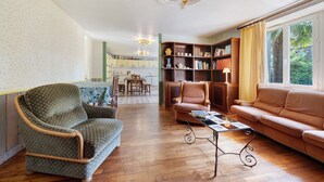 Un salon comprenant un canapé, un fauteuil et une table basse. On y trouve une bibliothèque remplie de divers objets, et un coin repas à l’arrière-plan.
