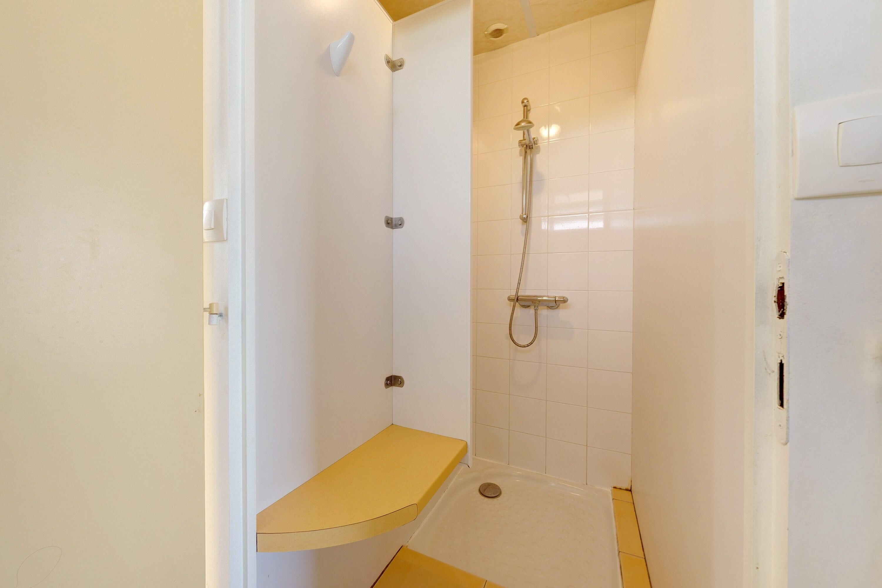 A small, enclosed shower area with a bench, a handheld showerhead, and a wall-mounted control panel.