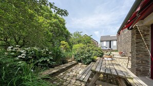 Une terrasse en pierre avec un banc en bois, entourée d’une végétation luxuriante et d’un bâtiment en pierre.