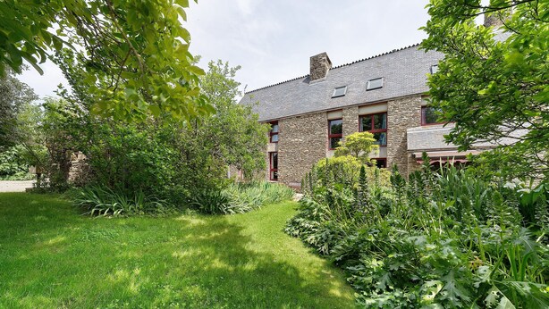 Un bâtiment en pierre doté d’un toit en pente, d’une cour verdoyante et d’un feuillage dense.