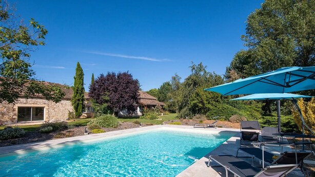 Une piscine entourée de verdure et de bâtiments en pierre, avec des chaises longues et des parasols.