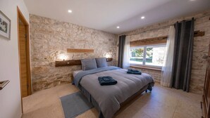Une chambre avec un mur en pierre, un lit recouvert d’une couverture grise, une table de chevet avec une lampe et une fenêtre avec des rideaux.