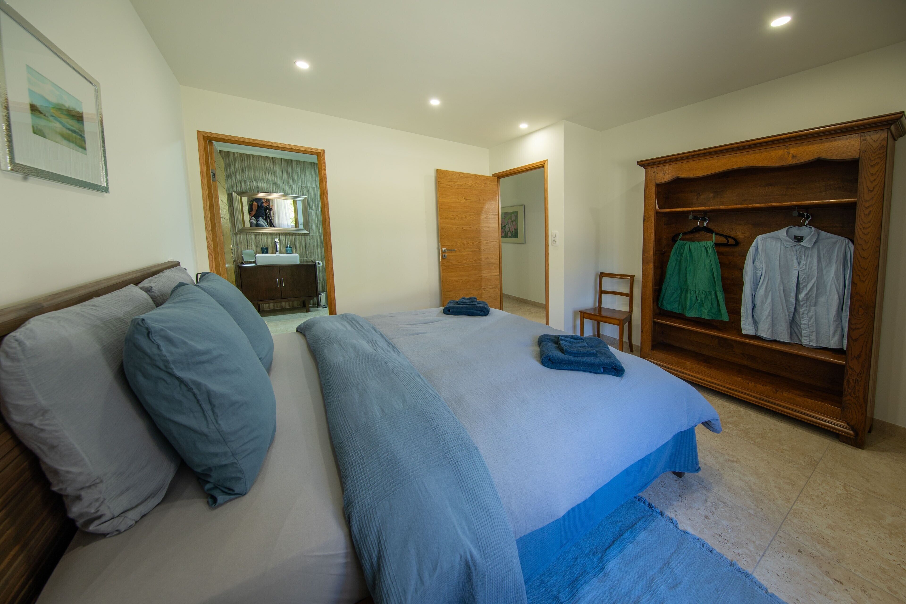 A bedroom with a bed, a wooden wardrobe, a mirror, and a bathroom visible through an open door.