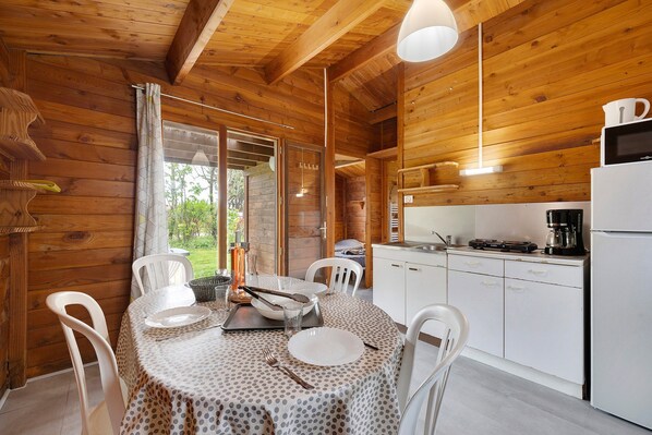 A wooden cabin with a dining table set for two, a kitchen with basic appliances, and a view of greenery outside.