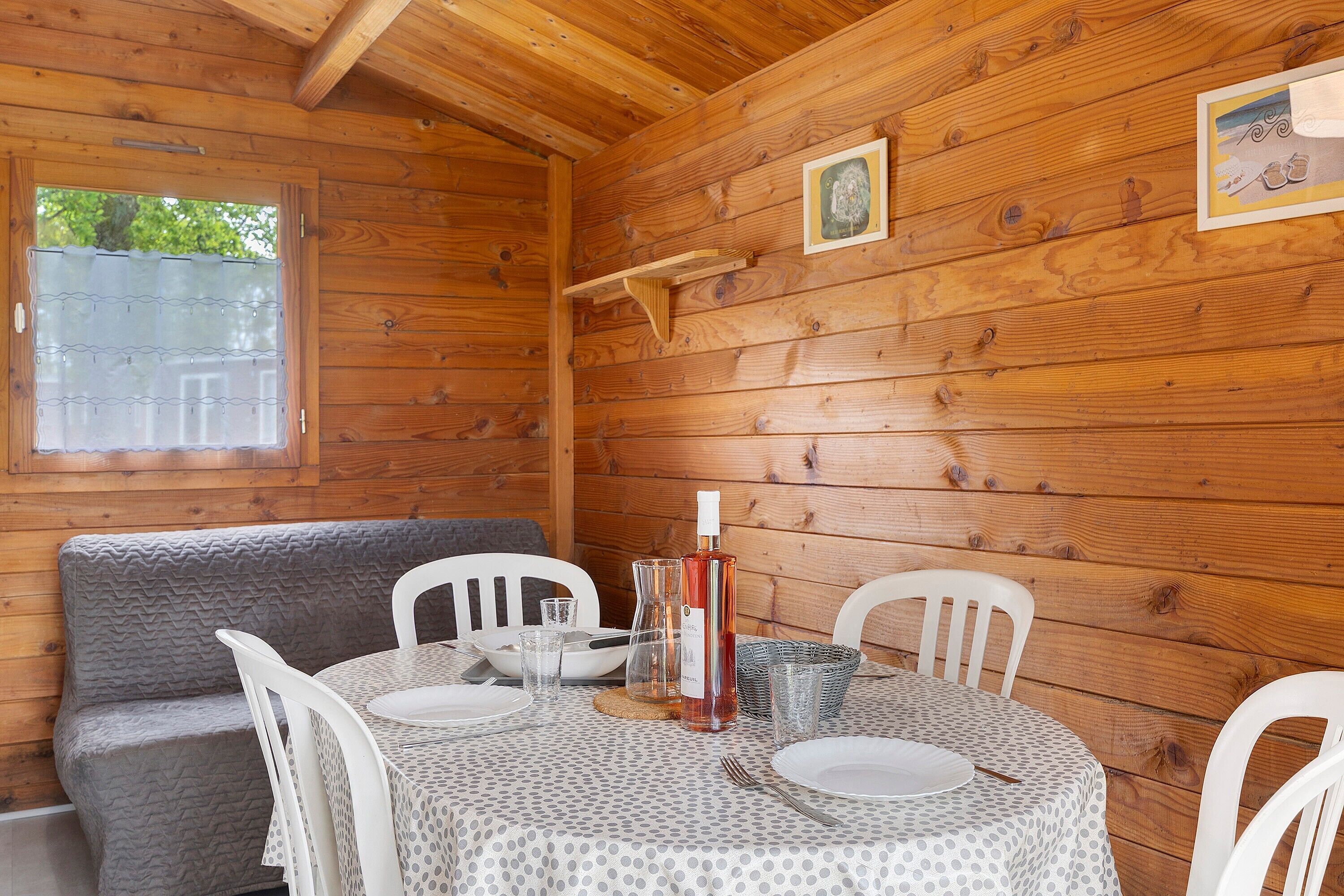 A wooden cabin interior with a round table set for dining, white chairs, a grey cushioned bench, and a wooden shelf with a picture.
